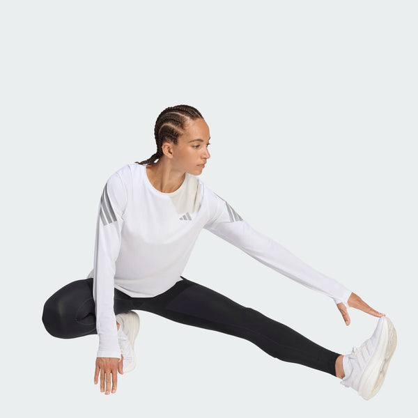 En person iført en Adidas Adi365 Long Sleeve Shirt, Dame, i hvid sidder på gulvet, strækker det ene ben ud til siden og rækker ud mod foden.