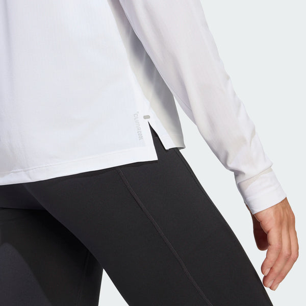 En kvinde ses fra taljen og ned iført sorte leggings og en Adidas Adi365 Long Sleeve Shirt i hvid, med den ene arm afslappet langs siden mod en almindelig, lys baggrund.