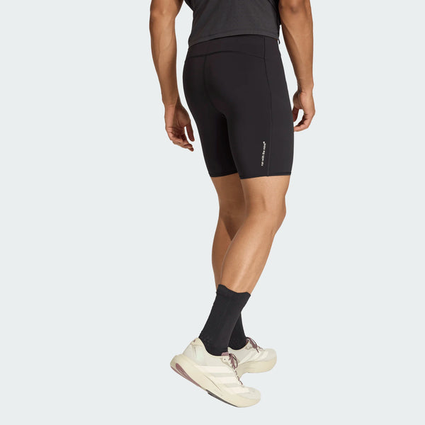 En person iført Adidas Adi365 Hk Tight shorts i sort står med ryggen til kameraet, parret med en mørk t-shirt, sorte strømper og hvide sportssko mod en almindelig lys baggrund.