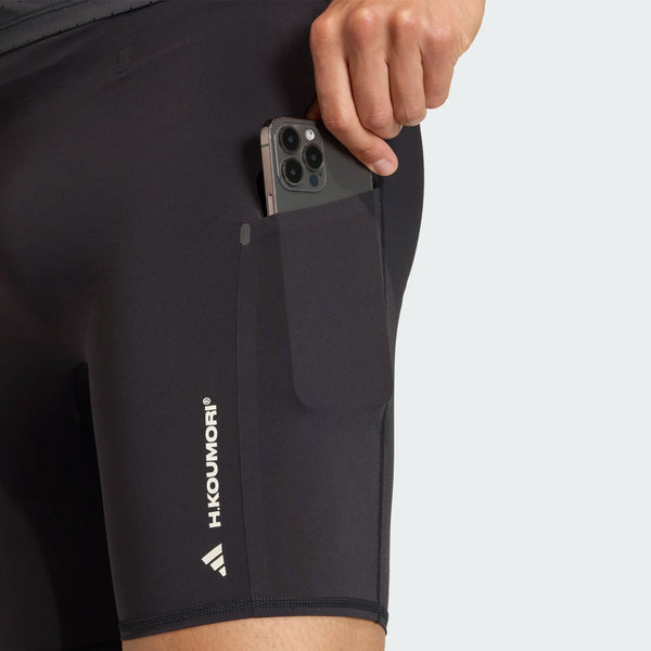En person putter en smartphone i sidelommen på sorte Adi365 Hk Tight shorts fra Adidas, som har mærkets navn trykt i hvidt på låret.