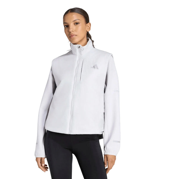 En person står med ansigtet fremad og har en lysegrå Adidas Adi365 Running Warm Vest til kvinder (dame) i farven Greone på, med høj krave og sort leggings. Vesten har et diskret Adidas-logo på venstre bryst og bæres over en langærmet bluse. Baggrunden er ensfarvet hvid.