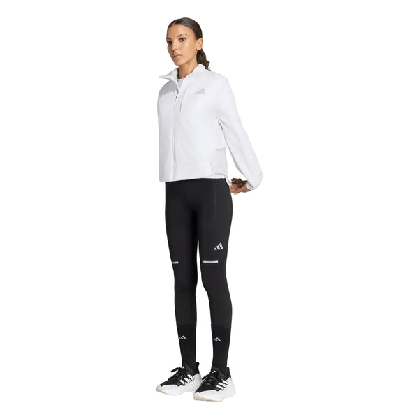 En person står med ansigtet vendt fremad og har en hvid sportsjakke med lynlås på, over hvilken der er taget en Adidas Adi365 Running Warm Vest til kvinder i grøn. Derudover bærer personen sorte leggings med et lille logo, sorte knæstrømper og sort-hvide sportssko, alt sammen foran en ensfarvet hvid baggrund.