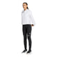 En person står med ansigtet vendt fremad og har en hvid sportsjakke med lynlås på, over hvilken der er taget en Adidas Adi365 Running Warm Vest til kvinder i grøn. Derudover bærer personen sorte leggings med et lille logo, sorte knæstrømper og sort-hvide sportssko, alt sammen foran en ensfarvet hvid baggrund.