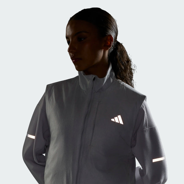 En person iført Adidas Adi365 Running Warm Vest til kvinder i farven Greone, med reflekterende striber på ærmerne og et reflekterende logo på brystet, står foran en ensfarvet lys baggrund, hvor ansigtet delvist er i skygge.