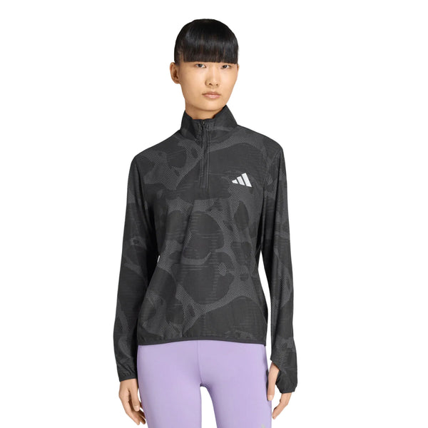 En person står med ansigtet fremad og har en Adidas Adi365 Running Half Zip-trøje til kvinder i farven Black/Grefiv på. Trøjen har et sort mønstret design med lange ærmer, en delvis lynlås foran, det hvide Adidas-logo og climawarm+-teknologi. Trøjen er kombineret med lyselilla leggings. Baggrunden er ensfarvet hvid.