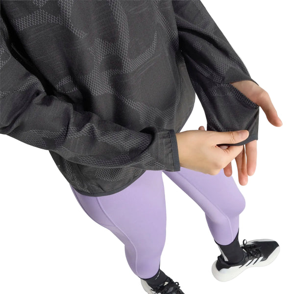 En kvinde ses iført en Adidas Adi365 Running Half Zip i farven Black/Grefiv, der har et mønstret design med lange ærmer og tommelfingerhuller. Hun har den på sammen med lyselilla leggings og sorte sportssko. Hendes ansigt kan ikke ses, da hun med højre hånd retter på trøjen med halv lynlås, hvilket fremhæver dens funktionelle design.