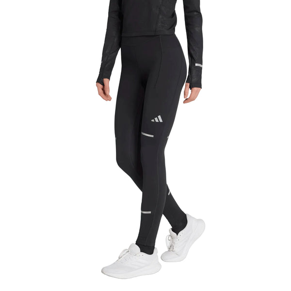 En person iført sorte Adidas Adi365 Climawarm+ løbetights (Dame) med reflekterende detaljer, en sort langærmet trøje og hvide sneakers står foran en ensfarvet hvid baggrund.