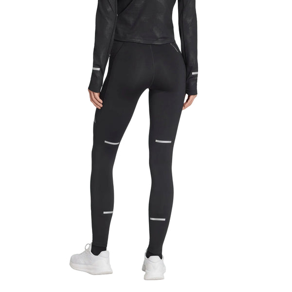 En person står med ryggen til kameraet og er iført en sort langærmet bluse, hvide sneakers og et par sorte Adidas Adi365 Climawarm+ løbetights til kvinder med diskrete refleksstriber. Baggrunden er ensfarvet hvid.