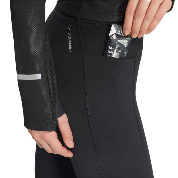 En person, hvor kun underkroppen og hånden er synlige, lægger en pakke med påskriften »Mautten Gel 100« i sidelommen på et par sorte Adidas Adi365 Climawarm+ løbetights (Dame), mens vedkommende har en langærmet trøje på.