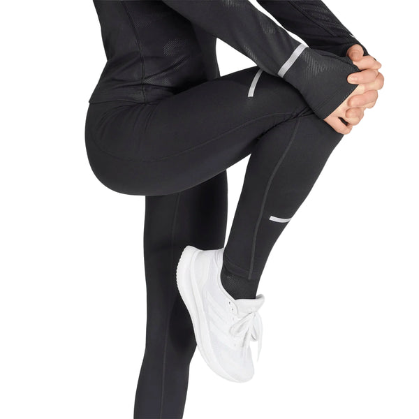 En person iført sorte Adidas Adi365 Climawarm+ løbetights (Dame) og en sort langærmet trøje samt hvide sneakers strækker sig ved at trække det ene knæ op mod brystet. Kun underkroppen og en del af den ene arm er synlige.
