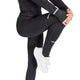 En person iført sorte Adidas Adi365 Climawarm+ løbetights (Dame) og en sort langærmet trøje samt hvide sneakers strækker sig ved at trække det ene knæ op mod brystet. Kun underkroppen og en del af den ene arm er synlige.