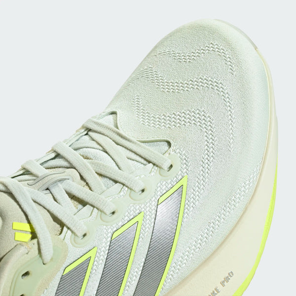 Et nærbillede af Adidas Supernova Prima 3, Herre i farvekombinationen Cryjad/Ironmt/Siggnr viser sølv- og gule striber, bølget syning på tåpartiet og et glimt af snøresystemet. Den cremefarvede sål har en neongul detalje langs kanten og tilbyder blød støddæmpning for overlegen komfort – hvilket gør den til et fremragende valg for mænd, der løber, og som værdsætter både stil og ydeevne.