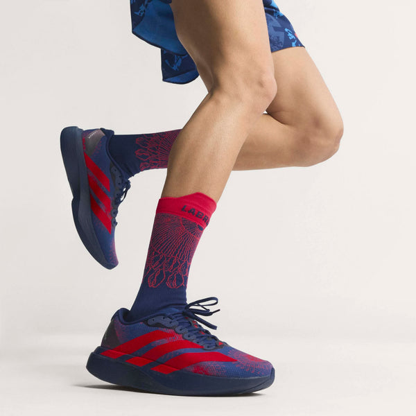 En person i Adidas Adizero Evo SL Woven løbesko herre, Dkblue/Lucred/Mutprp, med blå shorts og matchende sokker står med det ene ben let løftet mod en almindelig lys baggrund.