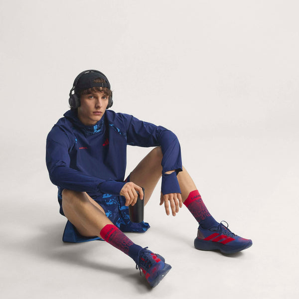 En person i blåt sportstøj, pink sokker, Adidas Adizero Evo SL Woven herre sko i Dkblue/Lucred/Mutprp og hovedtelefoner sidder på gulvet mod en hvid baggrund og holder en sort flaske.