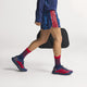 En mand går mod en almindelig baggrund iført blå og røde Adidas Adizero Evo SL Woven-sneakers (Herre, Dkblue/Lucred/Mutprp) med responsiv LIGHTSTRIKEPRO-støddæmpning, matchet med sportstøj og med en sort sportstaske i hånden.