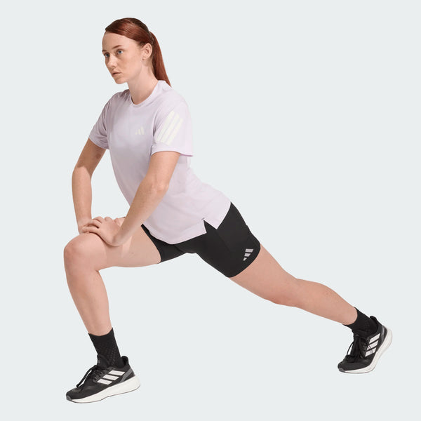 En rødhåret kvinde med hestehale laver en forlæns udstrækning iført Adidas Adi365 Running Tee, Dame, Icelav, sorte sportsshorts og -strømper og sorte sportssko med hvide striber på en almindelig hvid baggrund.