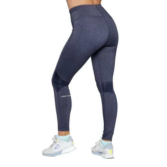 En person er iført mørkeblå Kari Traa Julie High Waist Tight, Dame, Marin leggings med KARI TRAA-logo på venstre underben og hvide sko med lyseblå og gule detaljer, stående på en hvid baggrund.