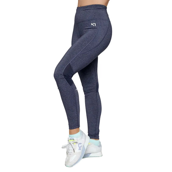 En person iført Kari Traa Julie High Waist Tight, Dame, Marin-høj talje og hurtigtørrende tights - og hvide sneakers med lyseblå og grønne detaljer - står på en almindelig hvid baggrund. Kun underkroppen og benene er synlige.