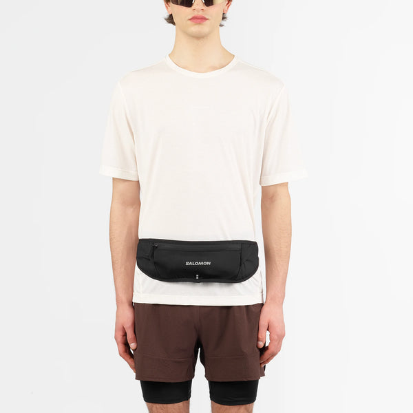 En person iført solbriller, en almindelig hvid t-shirt, brune shorts og Salomon Pulse Sling Belt (Unisex, sort) står på en almindelig hvid baggrund.