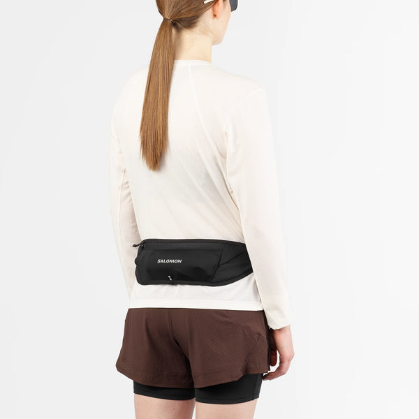 En person med langt hår, iført en hvid langærmet skjorte, brune shorts over sorte cykelshorts, et sort visir og Salomon Pulse Sling Belt (Unisex, sort), står med ansigtet vendt mod kameraet.