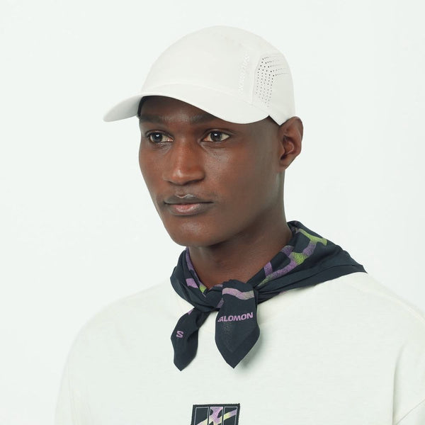 En person bærer en Salomon Shakeout Cap, Unisex, Whisper White, en hvid skjorte med en lille brystgrafik og et sort, grønt og lilla mønstret tørklæde bundet om halsen, stående mod en almindelig hvid baggrund.