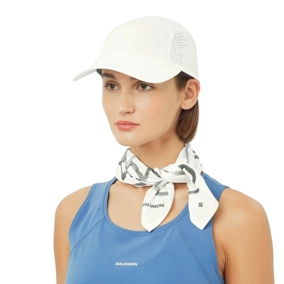 En kvinde iført en Salomon Shakeout Cap (Unisex, Whisper White), et hvidt tørklæde med et gråt mønster og en blå ærmeløs sportstop står på en almindelig hvid baggrund.