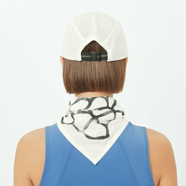 En person med kort brunt hår ses bagfra iført en Salomon Shakeout Cap i Whisper White baglæns, en blå ærmeløs top og en hvid bandana med et sort abstrakt mønster bundet om halsen.