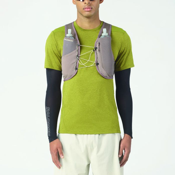 En person er iført en grøn kortærmet skjorte, hvide bukser, sorte ærmer og Salomon Gravel Skin 4 Set (Unisex, Iron/Silver Cloud) med vandflasker på brystet og fremviser innovativt løbeudstyr mod en almindelig hvid baggrund.