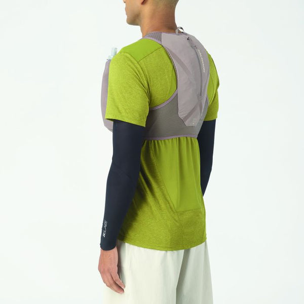 En person, der ses bagfra, er iført en lysegrøn T-shirt, hvide shorts, sorte ærmer og Salomon Gravel Skin 4 Set (Unisex, Iron/Silver Cloud), som skiller sig ud fra en almindelig hvid baggrund.