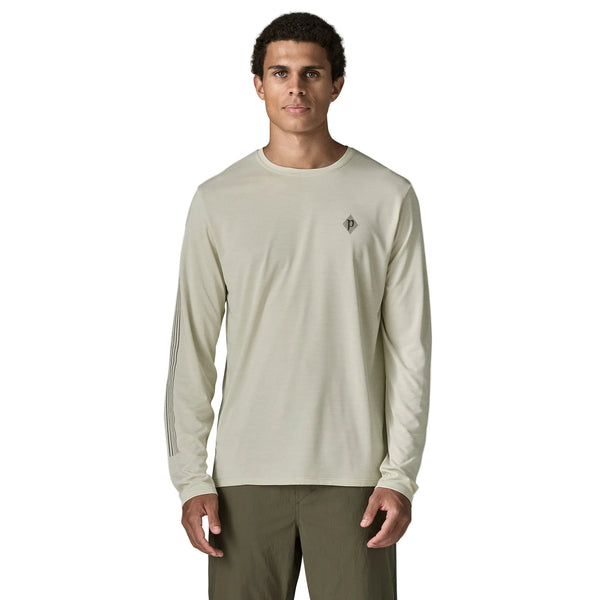 En mand står med ansigtet vendt fremad og har en lysbeige Patagonia L/S Cap Cool Daily Shirt – Hut Tripper, Herre, Dyno White – fremstillet af genbrugt polyester – samt olivengrønne bukser på. Hans arme hviler langs siden, og han har kort, krøllet hår. Baggrunden er ensfarvet hvid.