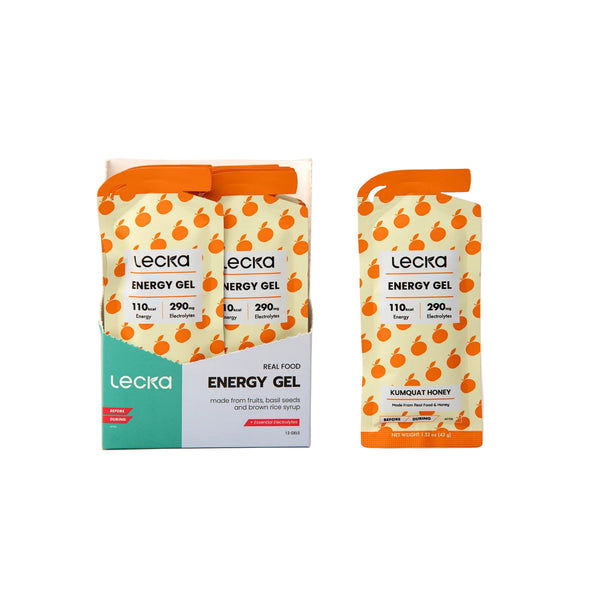 Her ses to Lecka Energy Gel, Kumquat/Honey-pakker og deres æske. De orange og hvide pakker har et frugtmønster, mens æsken viser Lecka-logoet, glutenfri-etiketten, indholdet af elektrolytter og produktoplysninger.