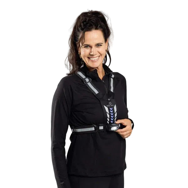En smilende kvinde har en sølvfarvet BeeSafe LED-sele med USB-telefonlomme, der lyser, på over en sort langærmet bluse.