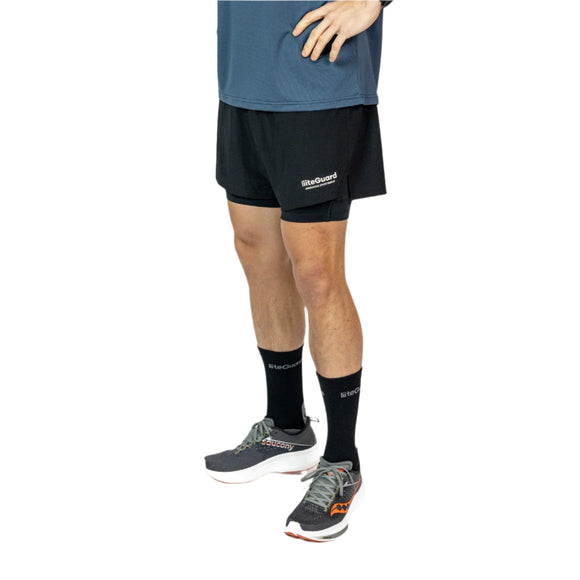 En person i blå skjorte, Liiteguard Glu-Tech Running 2In1 Shorts (Herre, Black), sorte sokker og grå løbesko står med den ene hånd på hoften. Billedet er brystbeskåret med en hvid baggrund.