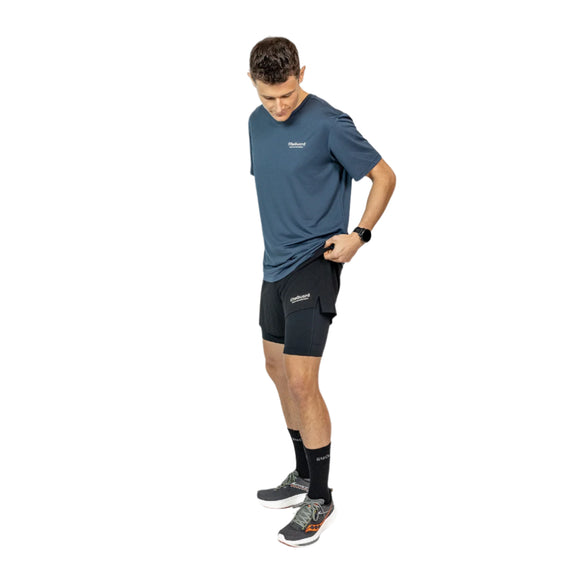 En mand er iført blå T-shirt, sorte strømper og Liiteguard Glu-Tech Running 2In1 Shorts, Herre, Black, mens han står på en hvid baggrund og kigger ned, mens han justerer linningen på sine shorts.