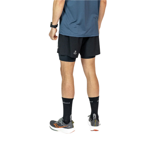 En person står med ryggen til og er iført Liiteguard Glu-Tech Running 2In1 Shorts til mænd i sort, høje sorte sokker, grå løbesko med orange detaljer og et sort ur. Kun den nederste halvdel af kroppen er synlig.