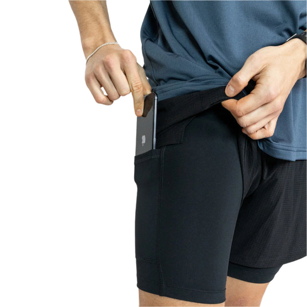 En person stikker en smartphone ned i sidelommen på Liiteguard Glu-Tech Running 2In1 Shorts, Herre, Black, mens han løfter sin blå skjorte en smule, så man kun ser overkroppen og overbenene mod en hvid baggrund. Ideelle herreshorts med Dri-FIT-teknologi.