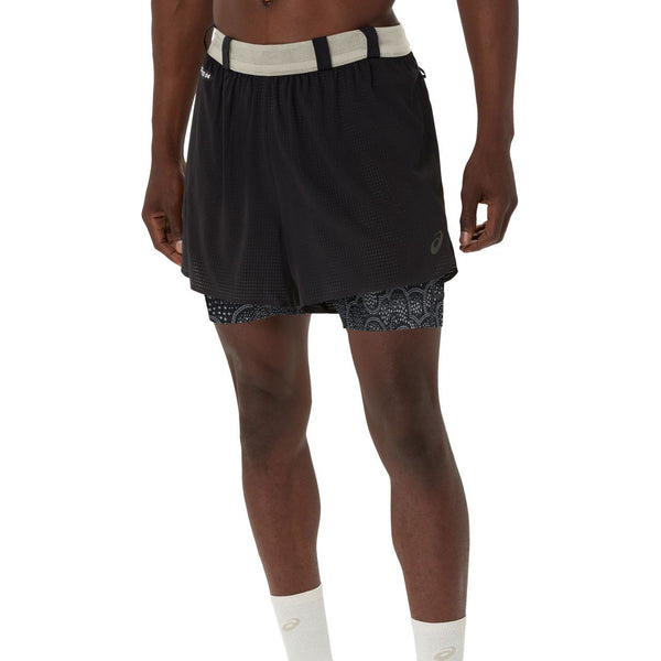 En person er iført ASICS Limited Series Metarun 4In 2-N-1 Shorts, Herre, i Performance Black/Cream med lyse mønstrede kompressionsshorts indenunder og hvide sokker. Shortsene har en elastisk linning og et lille logo på venstre ben.