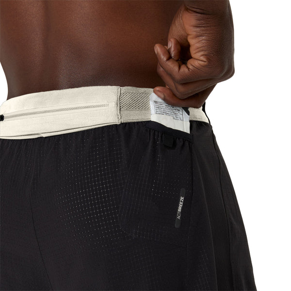 En person stikker en lille hvid genstand ned i baglommen på ASICS Limited Series Metarun 4In 2-N-1 Shorts, Herre, Performance Black/Cream, med let linning, diskret logo og synlige syninger.