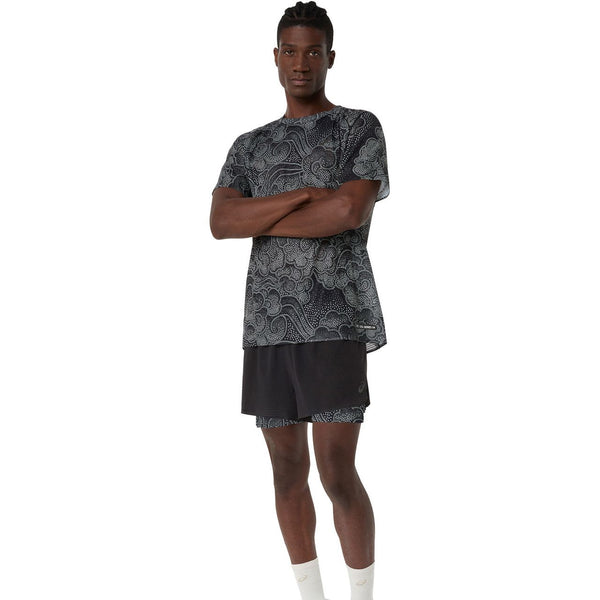 En person står med armene over kors iført en sort og hvid mønstret sportstrøje, Asics Limited Series Metarun 4In 2-N-1 Short til mænd i Performance Black/Cream og hvide sokker på en almindelig hvid baggrund.