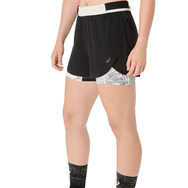 En person er iført Asics Limited Series Metarun Split Short (Dame, Performance Black) med hvid linning og mønstrede hvide kompressionsshorts indenunder, sammen med sorte mønstrede sokker og stående hænder afslappet i siden.