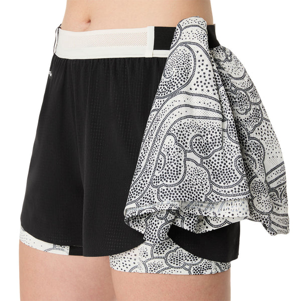 En person er iført Asics Limited Series Metarun Split Short, Dame, Performance Black, med en sort og hvid mønstret klud stoppet ind i linningen; det beskårne billede fokuserer på underkroppen og overbenene.