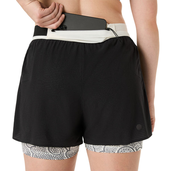 En person lægger en smartphone i baglommen på Asics Limited Series Metarun Split Short, Dame, Performance Black, med et hvidmønstret underlag, der titter frem forneden.