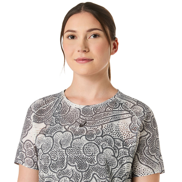 En kvinde med langt brunt hår bundet tilbage bærer ASICS Limited Series Metarun SS Top, Dame, Cream - med et sort abstrakt prikket mønster. Hun kigger fremad med et neutralt udtryk mod en almindelig hvid baggrund.