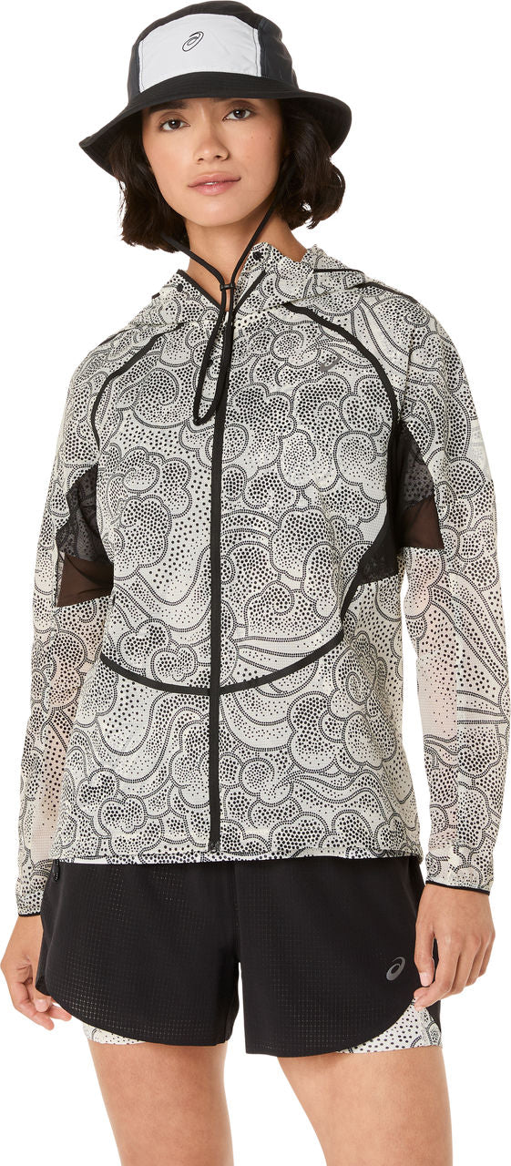 En person iført en ASICS Limited Series Wind Breaker Jacket til kvinder i cremefarvet står med front mod kameraet på en hvid baggrund. Hun kombinerer den med sorte shorts og en bucket hat, alt sammen med ASICS-branding.