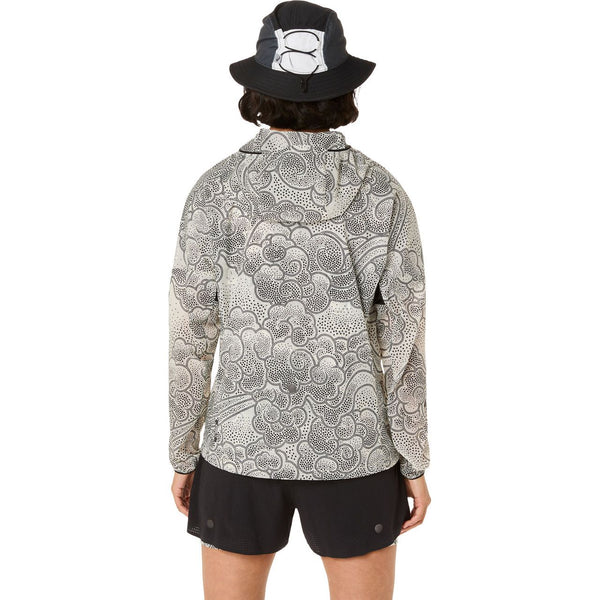 En person står og kigger væk fra kameraet iført en Asics Limited Series Wind Breaker Jacket, Dame, Cream med et sort og hvidt prikket design, parret med sorte shorts og en sort bucket hat med en hvid ryggrafik.