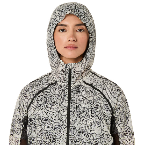 En kvinde kigger fremad i en cremefarvet Asics Limited Series Wind Breaker Jacket med et sort og hvidt abstrakt prikmønster. Jakken lukkes med lynlås, bæres med et neutralt udtryk og giver forbedret komfort med ACTIBREEZE-teknologi.