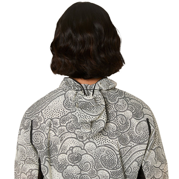 En person med bølget, skulderlangt mørkt hår ses bagfra iført en Asics Limited Series Wind Breaker Jacket, Dame, Cream, som har et sort-hvidt abstrakt prikket mønster på en cremefarvet baggrund.