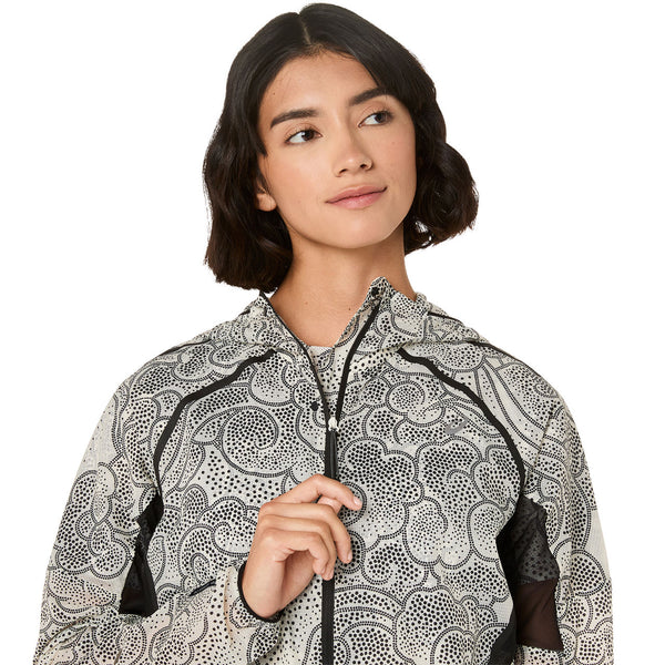 En person med kort mørkt hår bærer ASICS Limited Series Wind Breaker Jacket, Dame, Cream med et sort abstrakt prikket mønster, holder lynlåsen og kigger til siden mod en almindelig hvid baggrund.