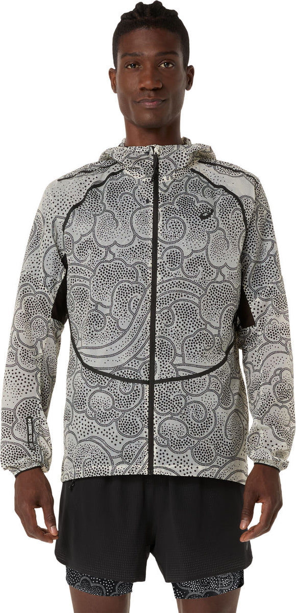 En person kigger fremad iført ASICS Limited Series Wind Breaker Jacket til mænd i Performance Black/Cream med sort abstrakt prikmønster, sort piping og sorte sportsshorts på en almindelig hvid baggrund.