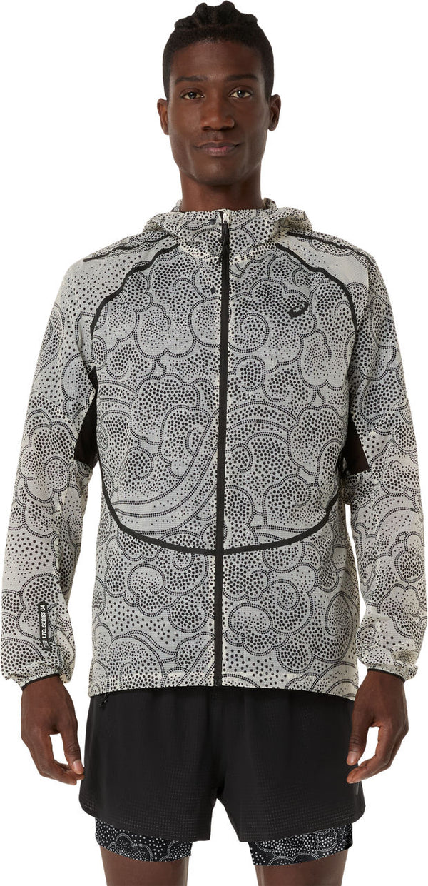 En person kigger fremad iført ASICS Limited Series Wind Breaker Jacket til mænd i Performance Black/Cream med sort abstrakt prikmønster, sort piping og sorte sportsshorts på en almindelig hvid baggrund.